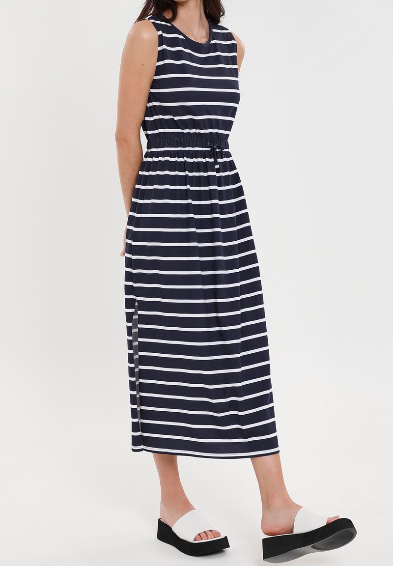 Robe maxi sans manches rayée bleu marine et blanche avec une taille élastique, des fentes sur les côtés, et associée à des sandales compensées blanches.