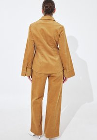 Woman with hair in a bun wearing mustard corduroy jacket and matching wide-leg pants, standing against a white background.
