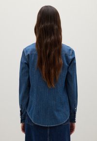 Camicia di jeans in blu medio con maniche lunghe, orlo curvo e polsini con bottoni. Il tessuto appare robusto con un design classico.