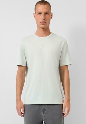 Mann mit kurzen Haaren, der ein einfaches, hellgraues T-Shirt mit kurzen Ärmeln und eine dunkelgraue Hose trägt, vor einem schlichten Hintergrund stehend.