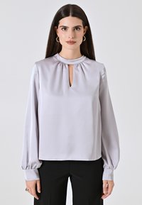 Blusa grigio chiaro in tessuto satinato, con scollo rotondo e un piccolo dettaglio a chiave, e maniche lunghe e voluminose.