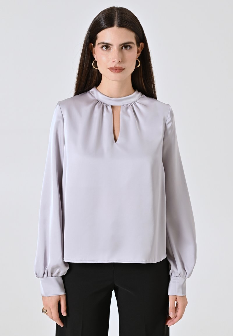 Blusa grigio chiaro in tessuto satinato, con scollo rotondo e un piccolo dettaglio a chiave, e maniche lunghe e voluminose.