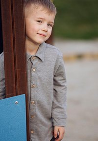 Szara koszula z długim rękawem z guzikami, stojąca obok brązowego słupa i niebieskiego panelu. Gładka faktura z delikatnymi detalami przeszyć.