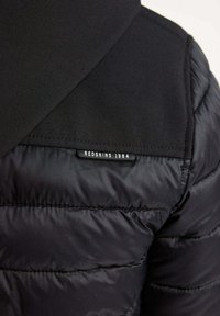 Doudoune noire avec une finition mate et un tissu texturé. Présente un design cousu et une étiquette indiquant "REDSKINS 1984" au dos.