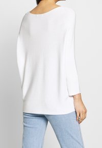 Femme portant un haut blanc ample à manches longues avec un col bateau et un jean bleu clair, vue de dos sur un fond uni.