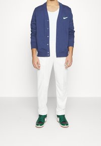 Cardigan blu navy con bottoni e logo Nike verde, abbinato a pantaloni bianchi e sneaker verdi con accenti neri e suole marroni.