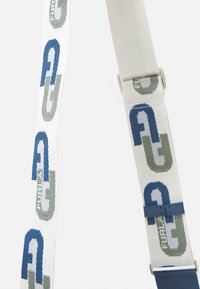 Vävt tygband i vitt, med blå och gröna geometriska logotyper. Inkluderar en blå läderdetalj och metalliska detaljer.