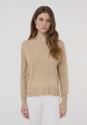 Maglione beige in maglia con maniche lunghe, scollatura rotonda e orlo e polsini a coste. Texture morbida e vestibilità rilassata. Indossato sopra pantaloni bianchi.