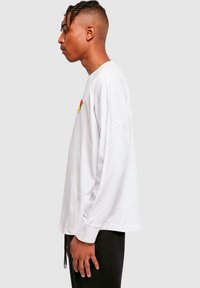 Hvid langærmet t-shirt med et farverigt grafisk design på brystet, afslappet pasform og ribbede manchetter, parret med sorte bukser.