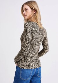 Vrouw met blond haar die een lange mouwen tijgerprint top en blauwe jeans draagt, zijwaarts poseert met handen in de zakken tegen een effen achtergrond.