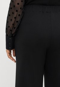 Czarne spodnie o gładkiej fakturze, z dopasowanym pasem i szerokimi nogawkami. Koszula ma przezroczyste czarne rękawy w kropki oraz guziki przy mankietach.