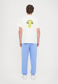 T-shirt en coton blanc avec un grand graphique de brocoli vert au dos, porté avec un pantalon de survêtement bleu clair et des baskets blanches.