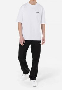 Man i vit t-shirt med kort ärm som har texten "PEGADOR" och svarta sweatpants med texten "PEGADOR", som går i vita sneakers.
