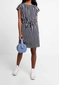Navy and white striped dress with a knotted waist, short sleeves, paired with a round blue handbag and white sneakers.
