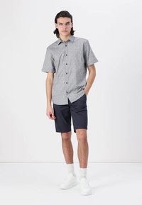 Cămașă cu mânecă scurtă în gri deschis, cu un model geometric, combinată cu pantaloni șort bleumarin și adidași albi. Butoane clare și croială casual.