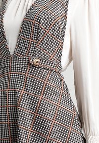 Yhdistelmä mustavalkoista houndstooth-kuviota, jossa oranssit ruudukkoaksentit. Mallissa on syvä V-pääntie ja suuri nappikoriste.
