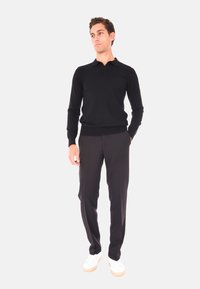 Uomo in piedi con le mani in tasca, che indossa una maglietta nera a maniche lunghe, pantaloni neri e sneaker bianche su uno sfondo bianco uniforme.