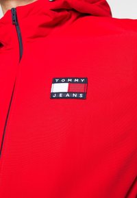 Rød hættejakke lavet af glat stof, med et sort og hvidt patch med "TOMMY JEANS" tekst og lagdelte farveblokke. Lynlåsdetalje synlig.