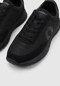 Zapatillas deportivas negras con un material textil suave y suela de goma. Presentan cordones, acentos tonales y un logo circular en el lateral.