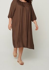 Robe marron jusqu'aux genoux avec encolure froncée et manches trois-quarts. Portée avec des sandales à lanières beige clair. Tissu lisse et fluide.