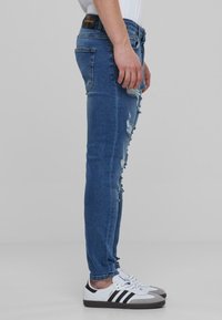 Kék skinny farmernadrág, sérült részletekkel a combokon és a térdeken. Fehér sportcipővel párosítva, fekete csíkokkal és szürke részletekkel.