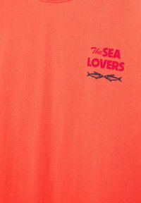 Koszula w kolorze koralowym z czerwonym napisem „The SEA LOVERS” oraz dwoma małymi czarnymi rybkami zwróconymi do siebie pod napisem na piersi.