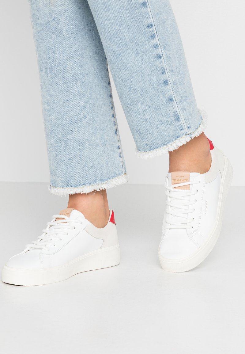 White leather sneakers with a textured surface, featuring a red heel accent and a beige tag on the tongue. Worn with light blue frayed jeans.