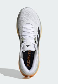 White athletic shoe with a textured knit upper, black stripes, green accents, and bright orange sole. Features flat laces and a padded collar.
