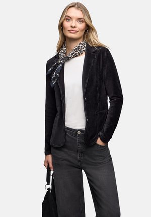 Sort kordjakke med en tætsiddende design, kombineret med en hvid top og mørke jeans. Et mønstret tørklæde tilføjer detaljer. Modellen holder en sort taske.