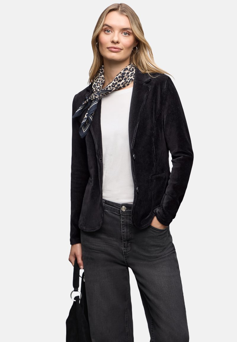 Czarna kurtka ze sznurkiem o dopasowanym kroju, zestawiona z białą bluzką i ciemnymi dżinsami. Wzorzysta chustka dodaje uroku. Modelka trzyma czarną torebkę.