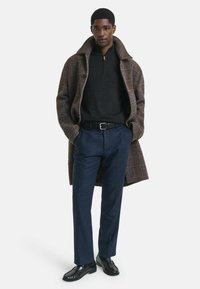 Bruin geruite overjas, zwarte ritsvest, marineblauwe broek en zwarte schoenen. De jas heeft een textuur en een strakke riemaccent.