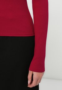 Top a maniche lunghe a coste in rosso scuro, con design aderente e polsini morbidi, abbinato a un fondo nero uniforme. Texture morbida visibile.