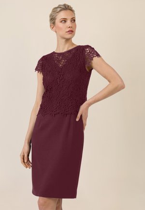 Frau in einem burgunderroten Kleid mit kurzem Spitzenoberteil und figurbetontem Rock, die mit einer Hand in der Hüfte vor einem schlichten beigefarbenen Hintergrund posiert.