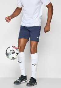 Shorts de sport bleus, t-shirt de sport blanc, chaussures de football noires et protège-tibias blancs. Le ballon de football présente un design noir et rouge.