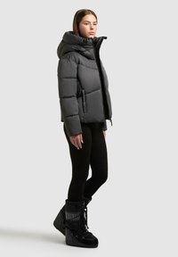 Giacca a vento nera con colletto alto e zip, caratterizzata da una forma squadrata e ampie sezioni trapuntate. Abbinata a leggings neri e stivali da neve.