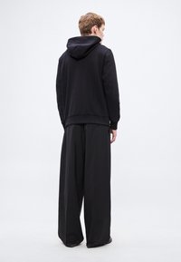 Μαύρο φούτερ με κουκούλα, με ριμπ μανσέτες και τελείωμα, σε συνδυασμό με φαρδιά μαύρη παντελόνια, που παρουσιάζει έναν μινιμαλιστικό σχεδιασμό και ματ υφή.
