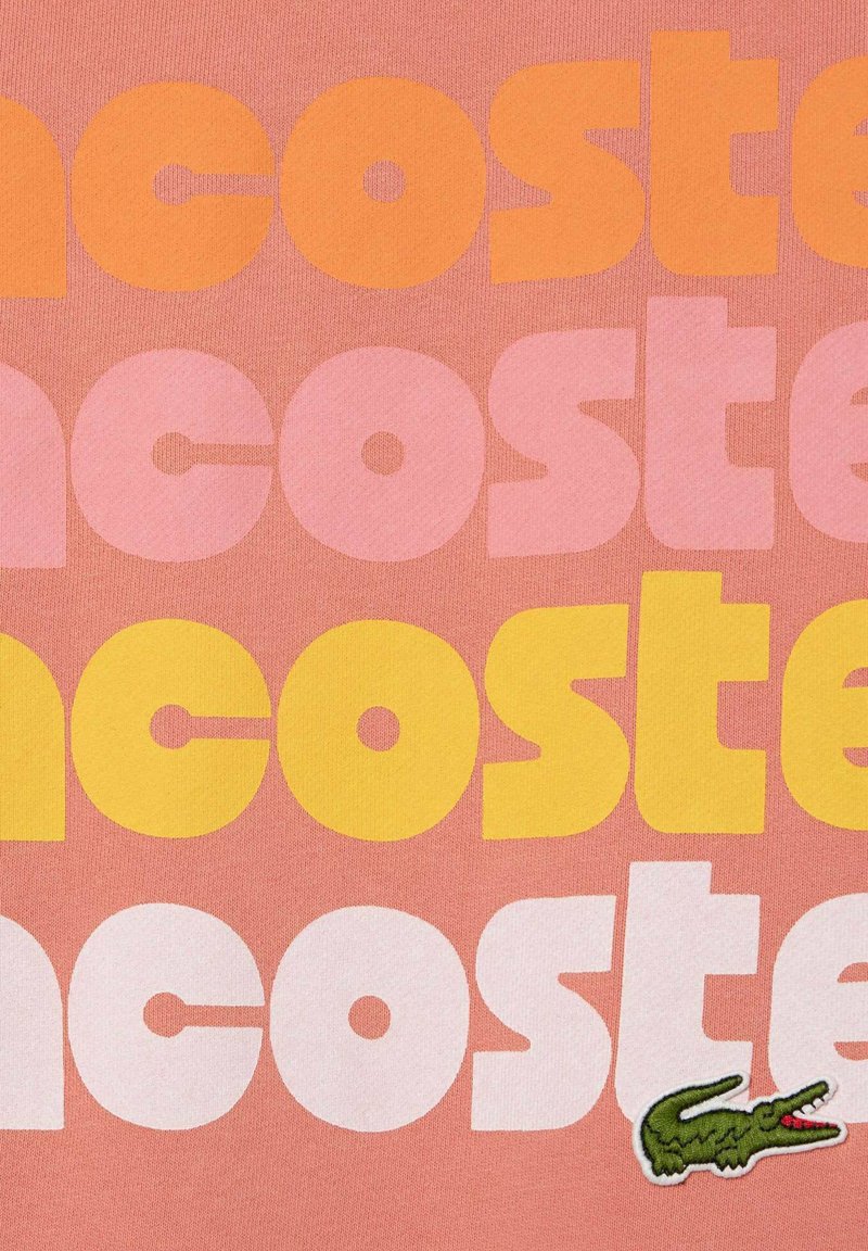 T-shirt rosa a maniche corte con grande testo sovrapposto in arancione, giallo e rosa, e logo di un coccodrillo ricamato in verde sul lato.