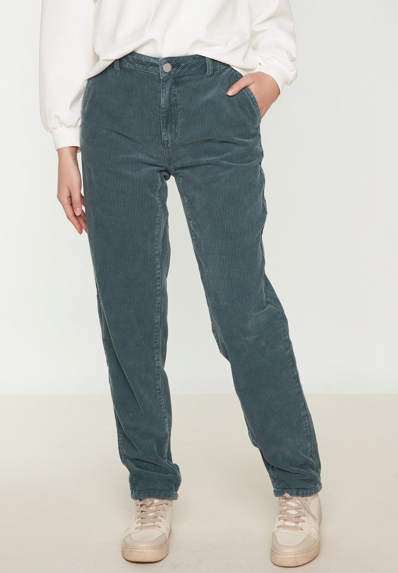 Personne portant un pantalon en velours côtelé bleu sarcelle, des baskets blanches et un haut blanc à manches longues, debout avec une main dans la poche.