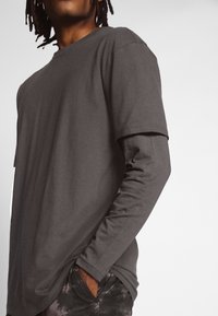 Gray long-sleeve shirt with a round neckline, soft fabric, slightly loose fit, and unfinished hems, paired with dark patterned pants.