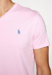 Ljusrosa bomulls T-shirt med V-urringning som har en liten blå broderad logotyp på vänster bröst. Kortärmad med en mjuk yta.