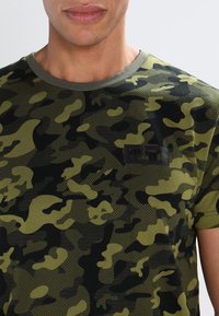 Grön camouflage-mönstrad T-shirt i mjukt tyg, med rund halsringning och en liten svart logopatch på bröstet.