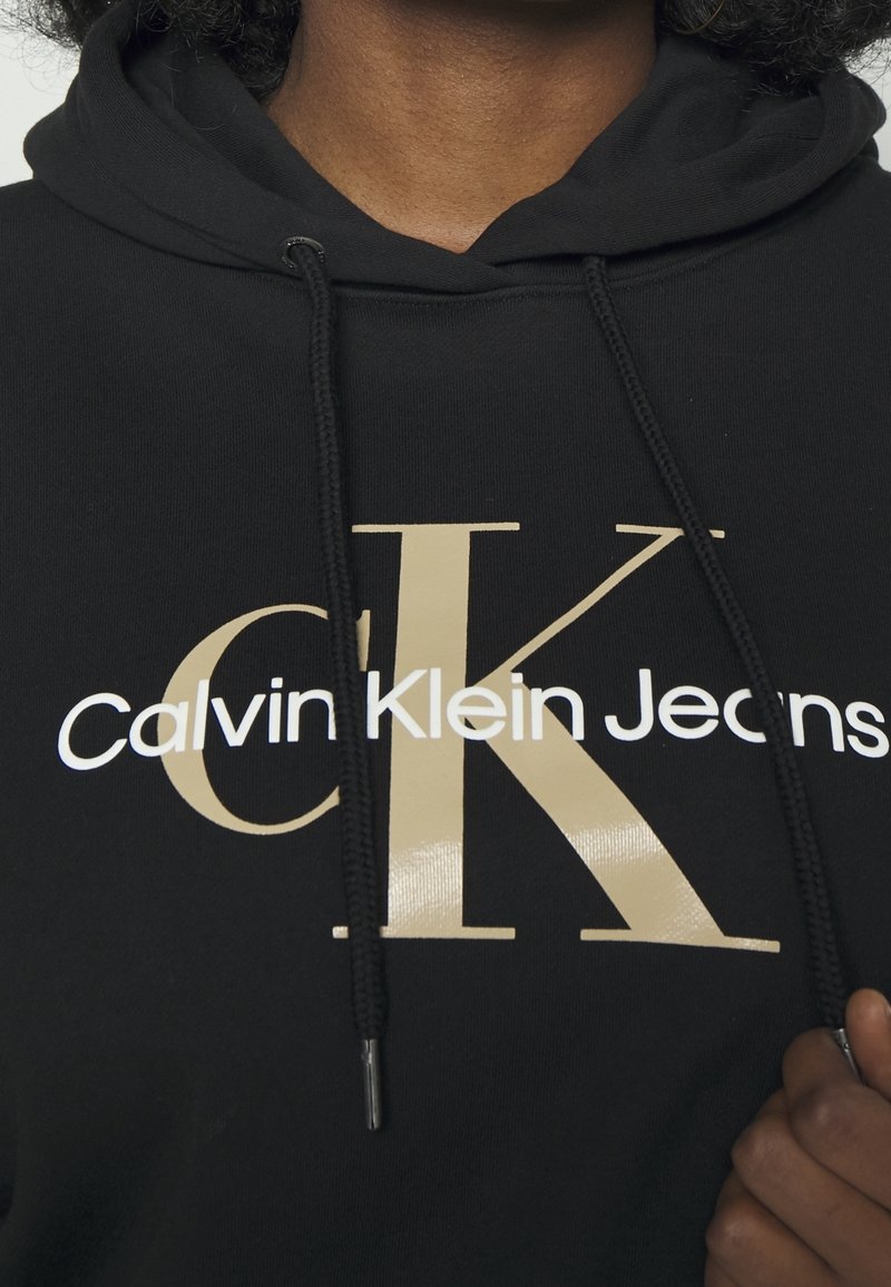 Svart hoodie med en guld- och vit Calvin Klein Jeans-logotyp. Har en huva med dragsko och en mjuk bomullstextur.