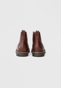 Par de botas de cuero marrón hasta el tobillo con tiradores y suelas negras resistentes, mostradas desde la parte trasera sobre un fondo blanco liso.