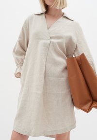 Beige vászonruha V-nyakkal, hosszú ujjakkal, elasztikus mandzsettákkal és laza fazonnal. Barna bőr tote táskával kiegészítve.