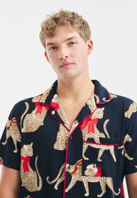 Koszulka nocna z krótkim rękawem, charakteryzująca się granatową bazą z wzorem lamparta, czerwonymi akcentami oraz świątecznymi detalami, takimi jak czapki Mikołaja i wstążki.