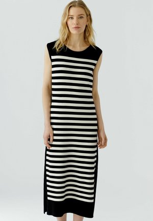Femme portant une robe midi sans manches à rayures horizontales noires et blanches, debout devant un fond clair uni.