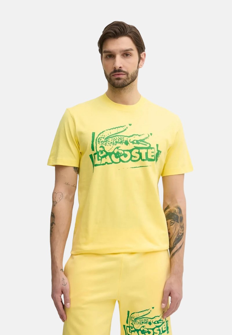 T-shirt di cotone gialla con logo di coccodrillo verde e "LACOSTE" stampato in verde. Maniche corte e scollatura a girocollo classica.