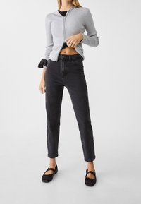 Cárdigan gris acanalado con cremallera y capucha, combinado con jeans de mezclilla negros de talle alto. Zapatos planos negros con correas cruzadas completan el conjunto.