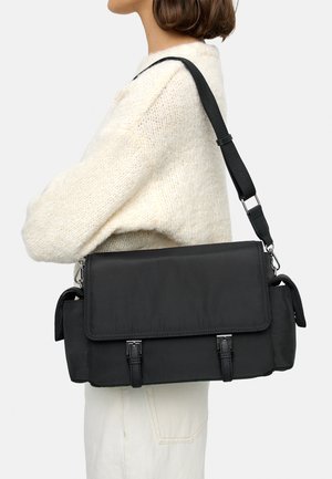 Osoba nosząca kremowy sweter z dzianiny i jasne spodnie, niosąca dużą czarną torbę listonoszkę z klapą zapinaną na klamry i regulowanym paskiem na ramię.