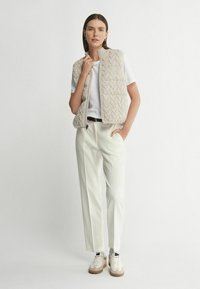 Beige Strickweste mit hohem Kragen, getragen über einem weißen T-Shirt. Kombiniert mit cremfarbigen Hosen und beigen Sneakern, die minimalistisches Design zeigen.